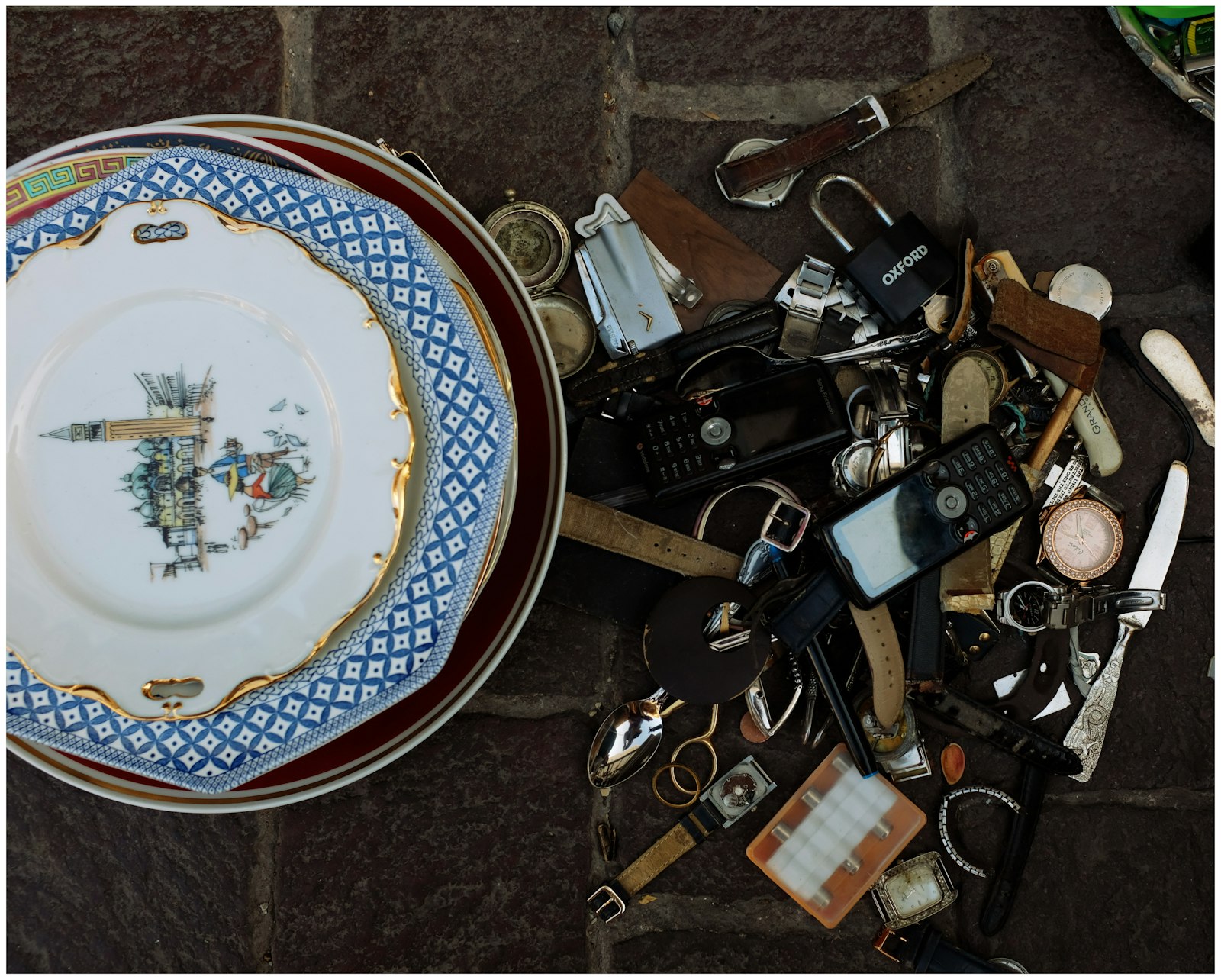 A flea market spread — porcelain plates, watches, locks, lighters laid out on stone — the kind of mixed inventory Margeen sorts through online.